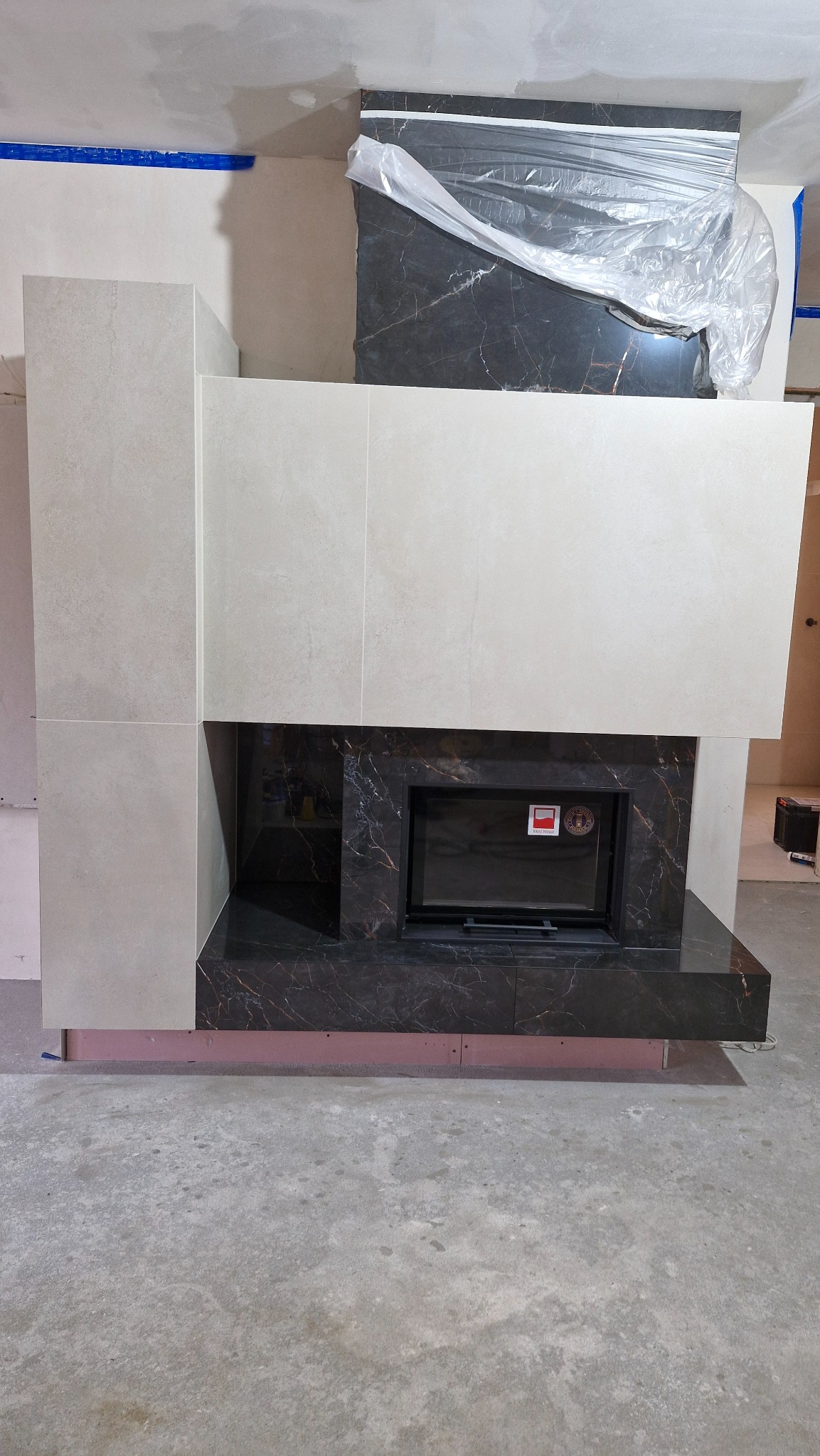 Obudowa kominka z płytek imitujących marmur i jasnego beżu, w trakcie montażu. Widoczne zabezpieczenia folią. Surowy stan podłoża, brak listew przypodłogowych.