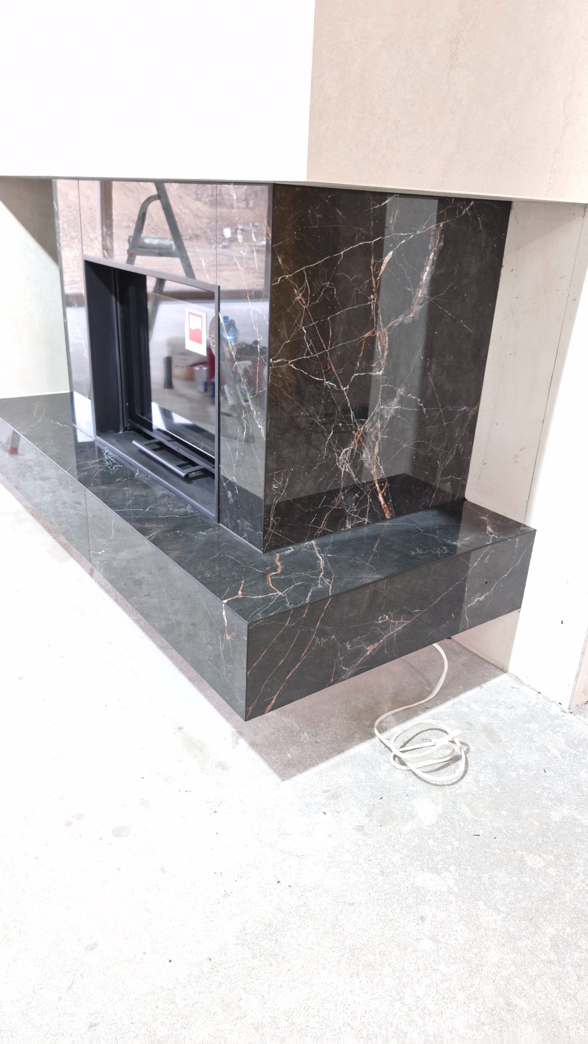 Wykończony kominek z czarnego marmuru z wyraźnym użyleniem, osadzony na podłodze w trakcie prac wykończeniowych. Widoczne przewody na posadzce.