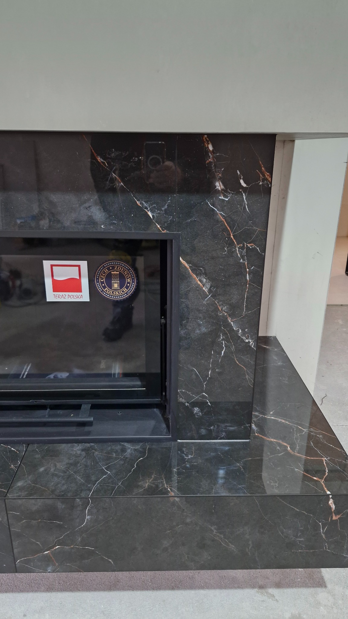 Obudowa kominka z czarnych płyt imitujących marmur z wyraźnym użyleniem. Widoczna szyba kominka z naklejkami 'Teraz Polska' i 'Produkt rekomendowany'.