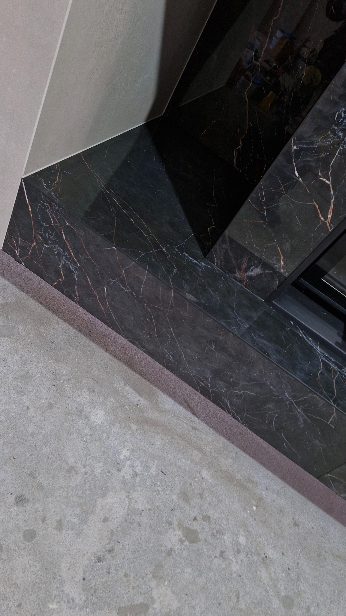 Fragment wykończenia wnętrza: czarne płytki marmurowe z czerwonymi żyłkami, szara ściana i surowa posadzka. Widoczne krawędzie i łączenia materiałów.