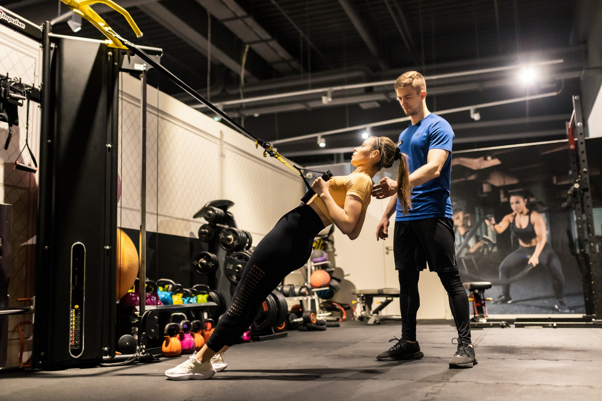 Kobieta ćwiczy na urządzeniu TRX w siłowni, z asekuracją mężczyzny, widoczne kettlebells w tle.