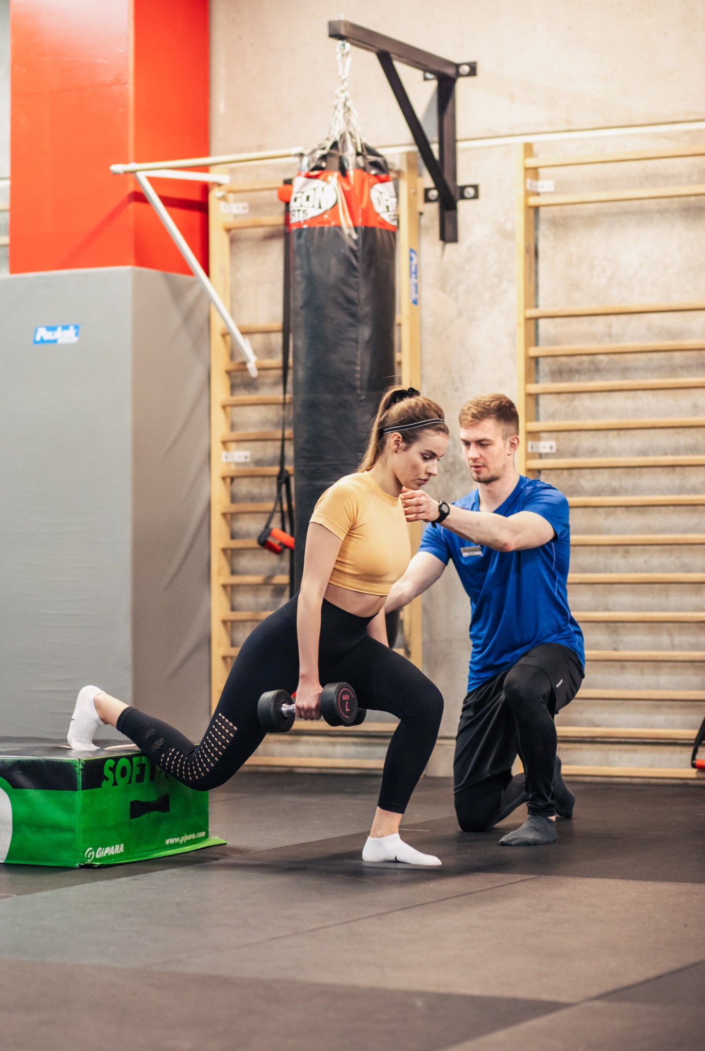 Kobieta w czarnych legginsach i żółtej koszulce wykonuje wypad w tył z hantlą na tle drabinek gimnastycznych, pod nadzorem mężczyzny w niebieskiej koszulce.