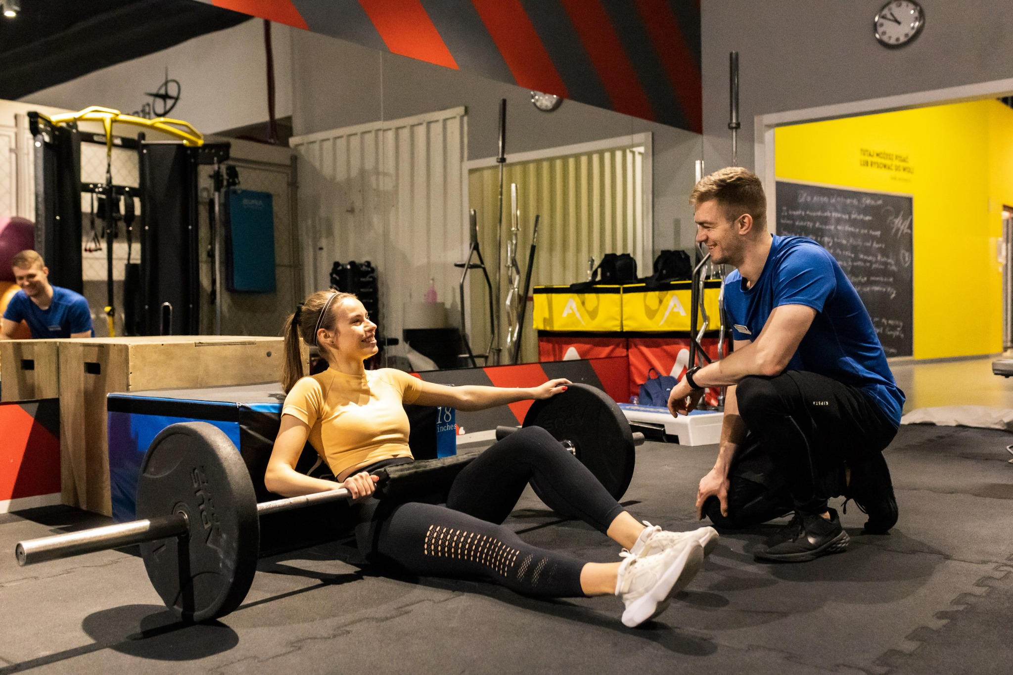 Kobieta w sportowym stroju wykonuje ćwiczenie z obciążeniem na biodrach, a uśmiechnięty mężczyzna w niebieskiej koszulce asekuruje, w tle widoczna siłownia z lustrem i sprzętem do ćwiczeń.