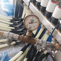 Instalatorstwo grzewcze LR LESKAROMA - Rozdzielacz instalacji grzewczej podłogowej z widocznym manometrem i rurami w izolacji, zainstalowany na folii budowlanej.