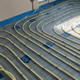 Instalatorstwo grzewcze LR LESKAROMA - Układanie rur systemu ogrzewania podłogowego na izolacji z siatką, widoczne mocowania rur do podłoża oraz fragment ściany i okna.