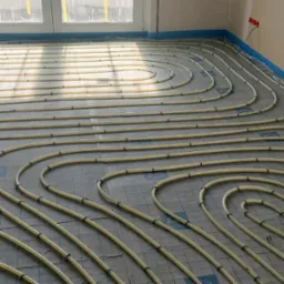 Układ rur grzewczych na podłodze, przygotowany do zalania wylewką, widoczne mocowania rur do podłoża i zabezpieczona folią ściana.