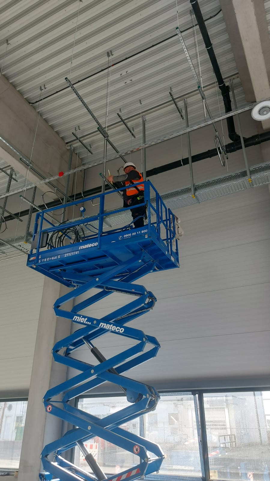 Pracownik w pomarańczowej kamizelce na podnośniku koszowym, montuje instalacje pod sufitem w hali przemysłowej. Widoczne rury i elementy mocujące.