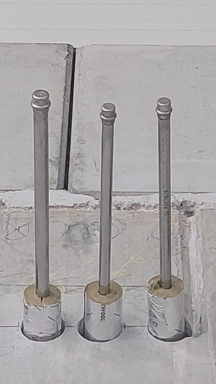 Trzy pionowe rury instalacyjne wystające z podłoża, zabezpieczone folią i izolacją, widoczne ślady po ołówku na podłożu.