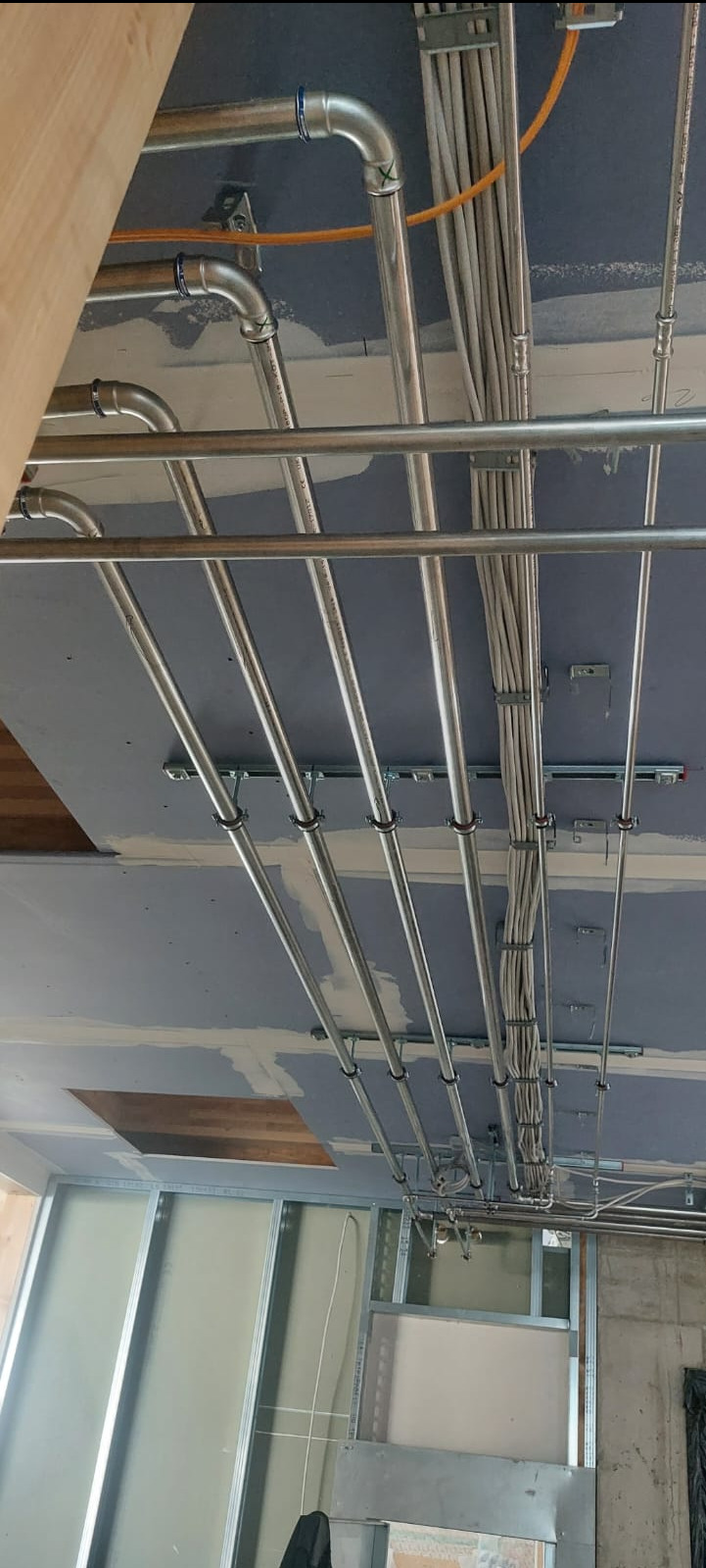 Instalacja rur i kabli pod sufitem podwieszanym w trakcie montażu, widoczne metalowe rury z niebieskimi złączkami i wiązki kabli przymocowane do konstrukcji sufitu.