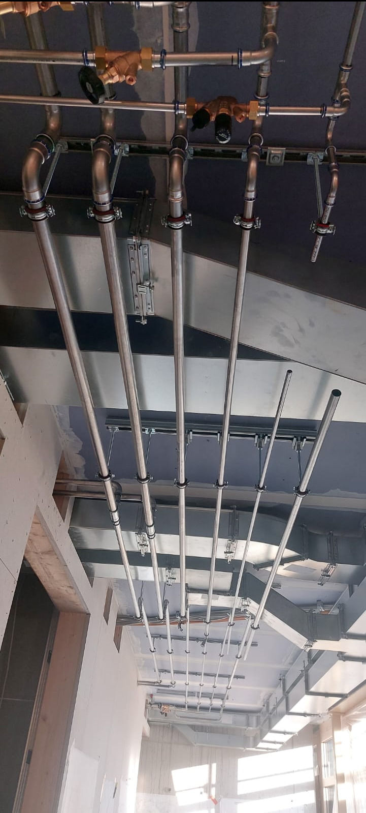 Widok z dołu na liczne rury instalacyjne, częściowo podwieszone, z widocznymi zaworami i mocowaniami, prowadzone pod sufitem w surowym stanie budynku.