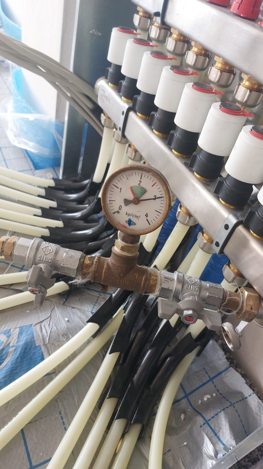 Rozdzielacz instalacji grzewczej podłogowej z widocznym manometrem i rurami w izolacji, zainstalowany na folii budowlanej.