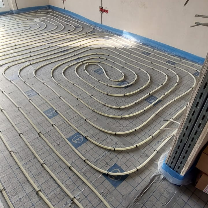 Układ rur w systemie rozprowadzania ciepła podłogowego, zamocowanych do izolacji podkładowej, widoczne uchwyty mocujące i przewody elektryczne w tle.