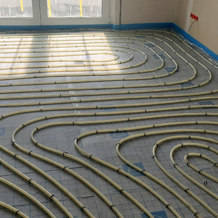 Układ rur grzewczych na podłodze, przygotowany do zalania wylewką, widoczne mocowania rur do podłoża i zabezpieczona folią ściana.