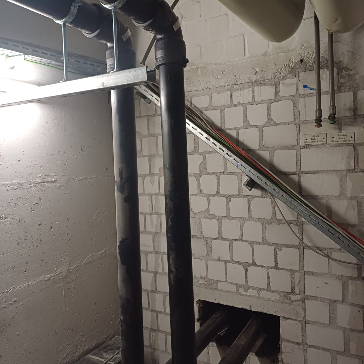 Fragment instalacji hydraulicznej z czarnymi rurami, widoczne mocowania do ściany z cegieł i kable w korytku.