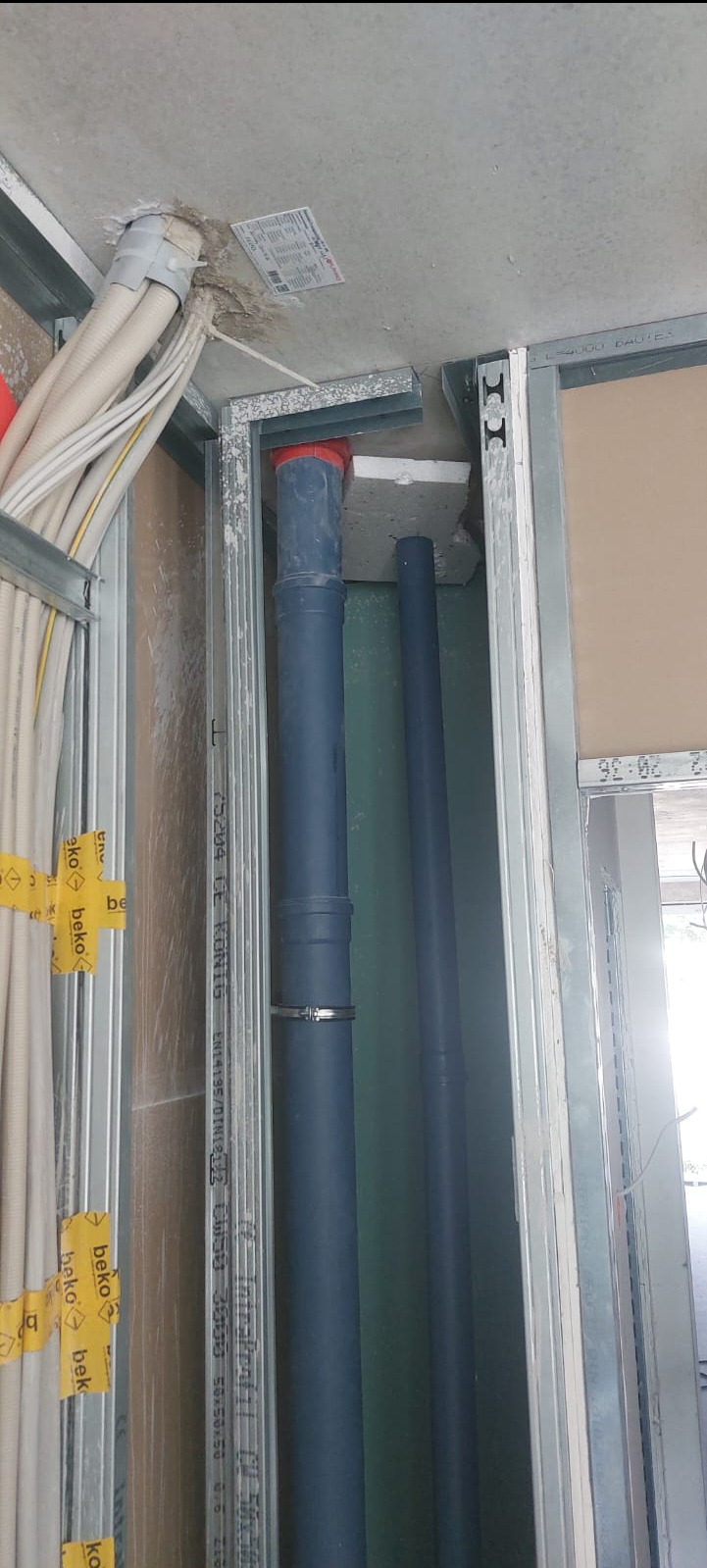 Dwie pionowe rury kanalizacyjne z tworzywa sztucznego, zamontowane w metalowej konstrukcji szkieletowej ściany, widoczne przewody elektryczne w peszlach i fragment płyty gipsowo-kartonowej.