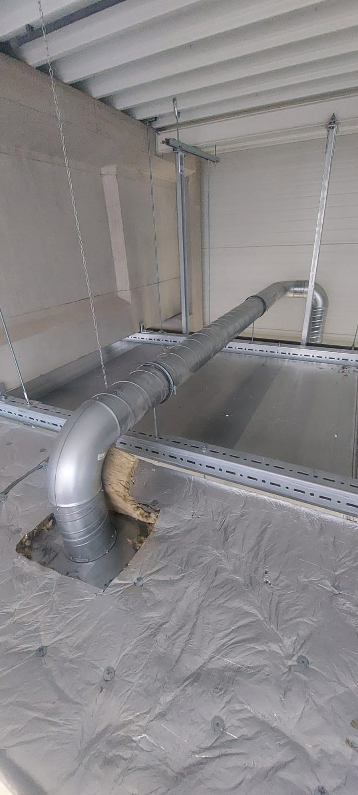 Srebrny kanał wentylacyjny z kolankiem, zamocowany pod sufitem na metalowych profilach, widoczna izolacja wokół przejścia przez podłogę.