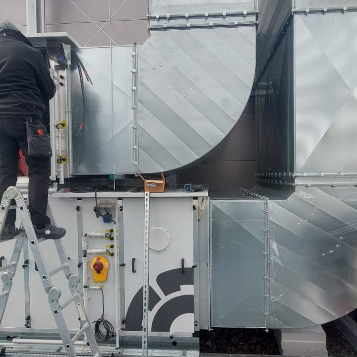 Pracownik na drabinie montuje system kanałów wentylacyjnych z blachy ocynkowanej, widoczny agregat z elementami sterującymi i logo producenta.