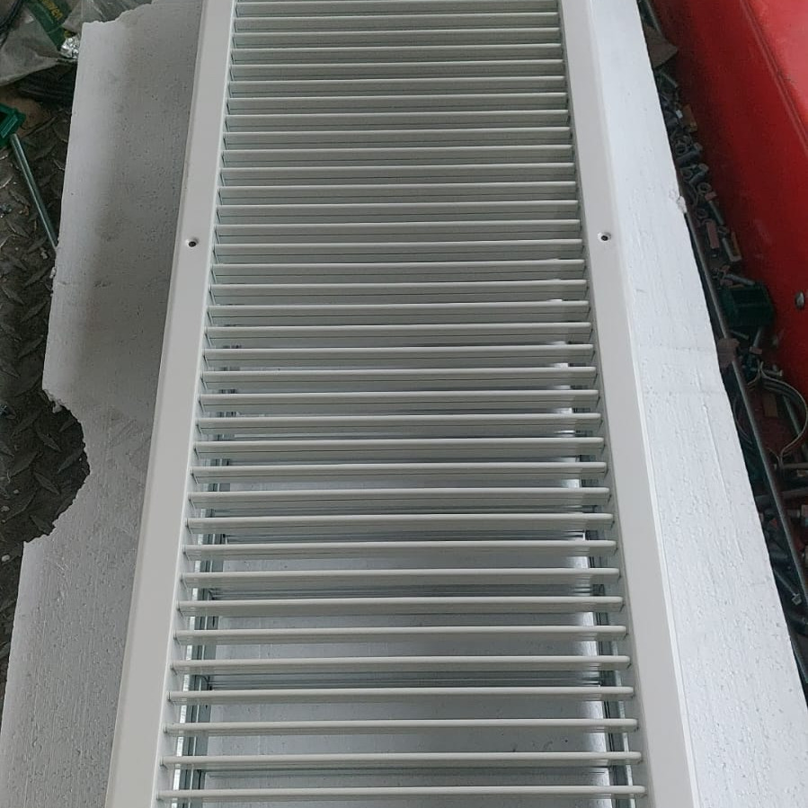 Biała, prostokątna kratka wentylacyjna z poziomymi szczelinami, leżąca na szarej, betonowej powierzchni. Widoczne ślady narzędzi i czerwony element w tle.