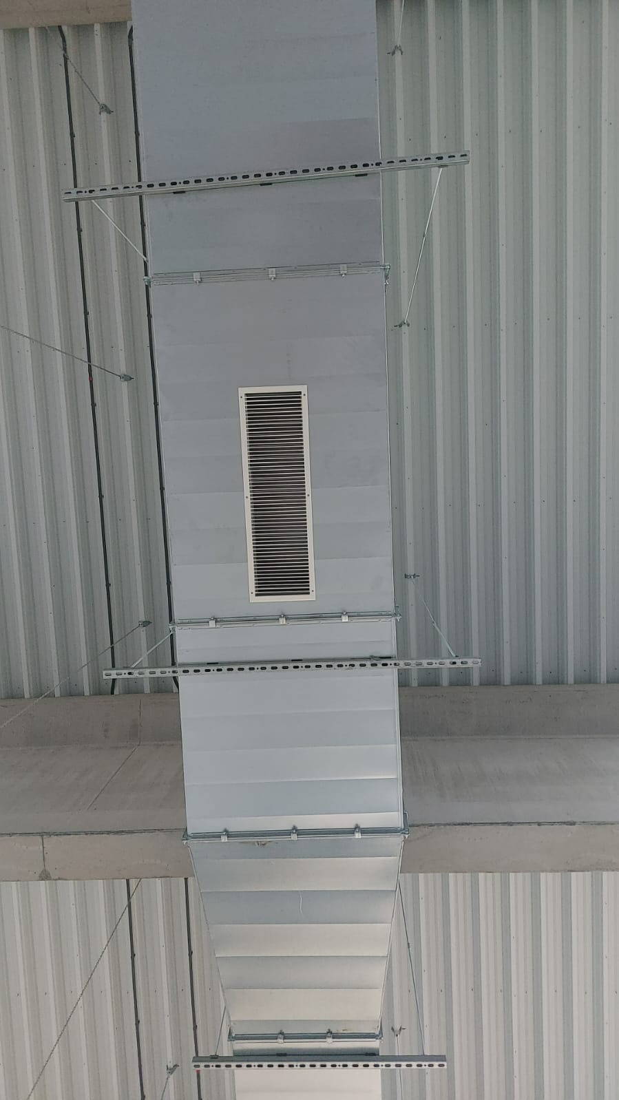 Szary kanał wentylacyjny prostokątny z kratką, zamocowany pod sufitem z blachy trapezowej, widok z dołu.