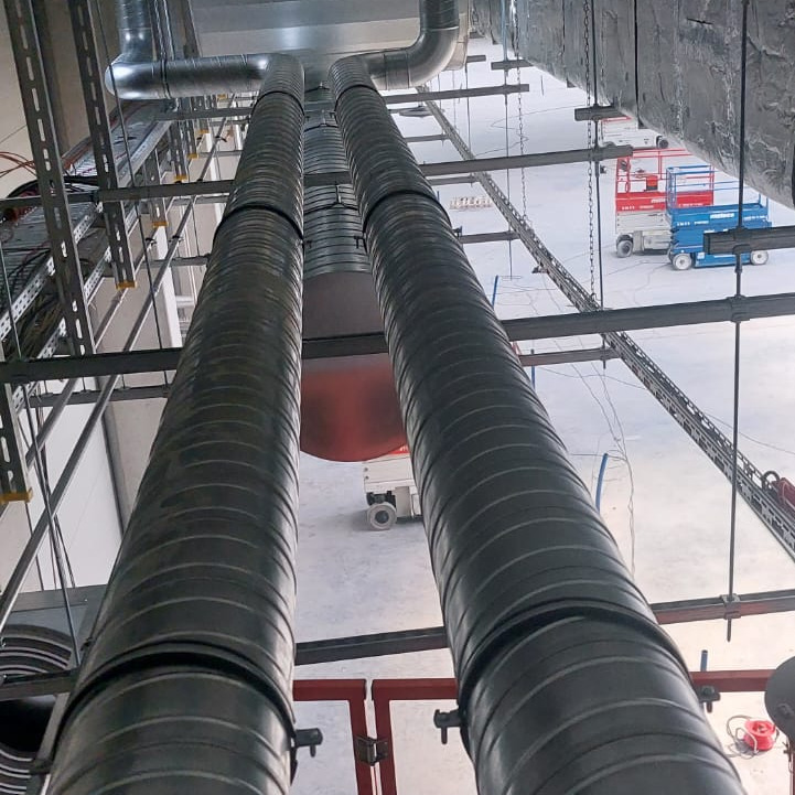 Dwa równoległe, spiralne przewody wentylacyjne z czarnymi opaskami, zamontowane pod sufitem hali, widoczne pod kątem, z maszynami podnośnikowymi w tle.