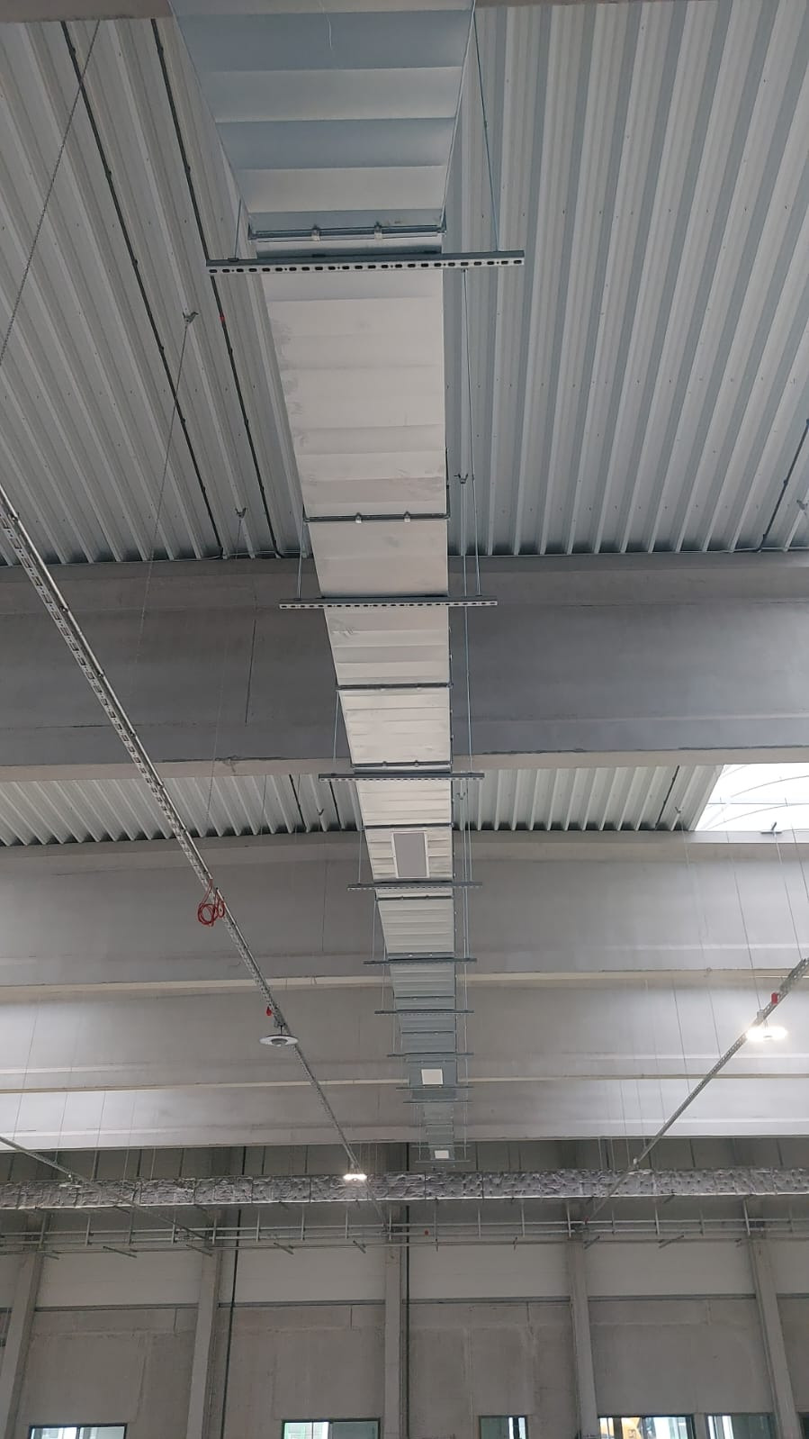Widok z dołu na system kanałów wentylacyjnych zamontowanych pod sufitem hali przemysłowej, widoczne elementy mocujące i oświetlenie.
