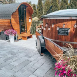 Mobilna sauna beczka z wejściem i balia z hydromasażem na przyczepie, ozdobione girlandą żarówek, z tabliczką informacyjną 'Mobilne Spa Kwiatek', otoczone zielenią i kwiatami w donicach.