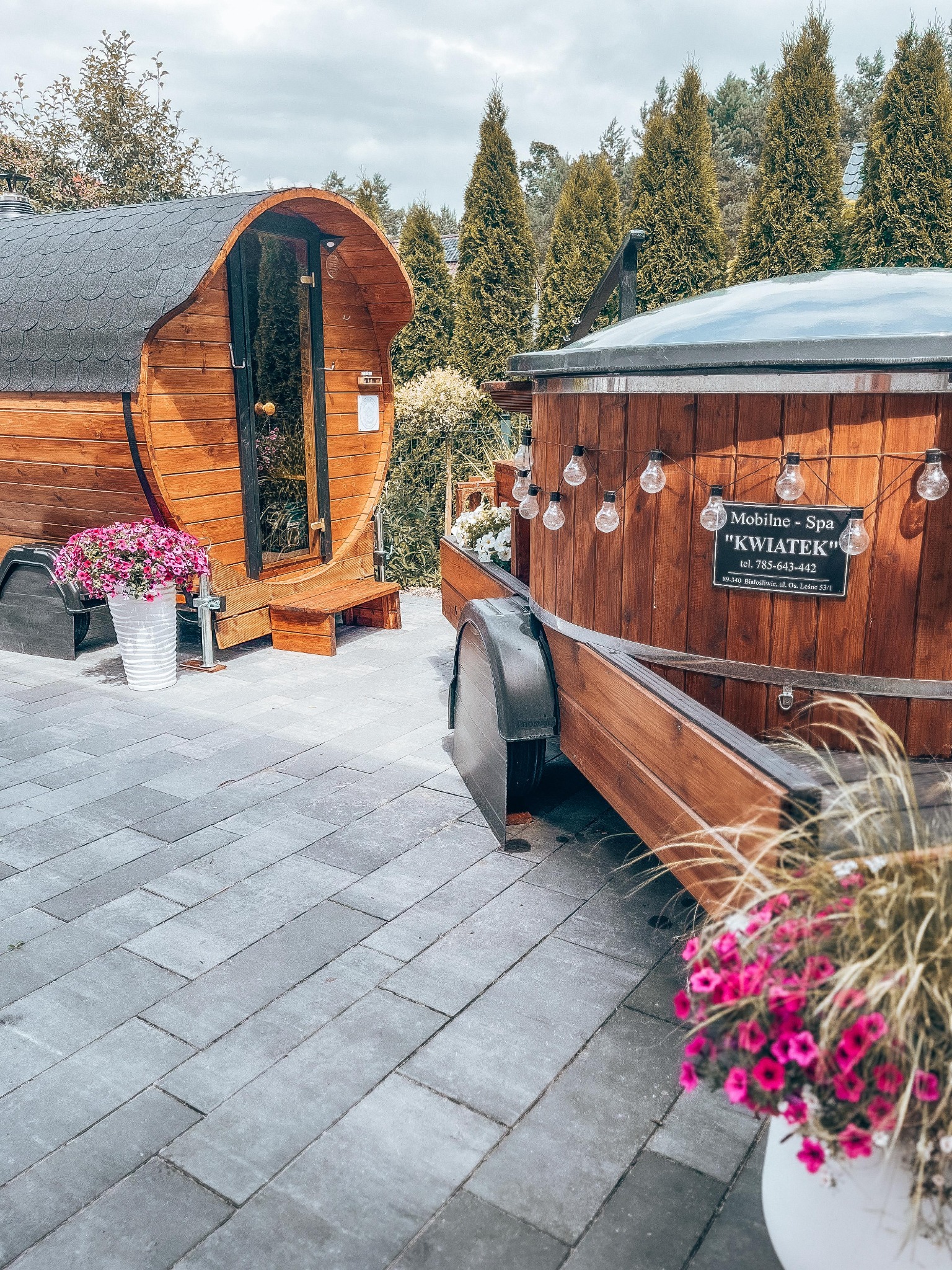 Mobilna sauna beczka z wejściem i balia z hydromasażem na przyczepie, ozdobione girlandą żarówek, z tabliczką informacyjną 'Mobilne Spa Kwiatek', otoczone zielenią i kwiatami w donicach.