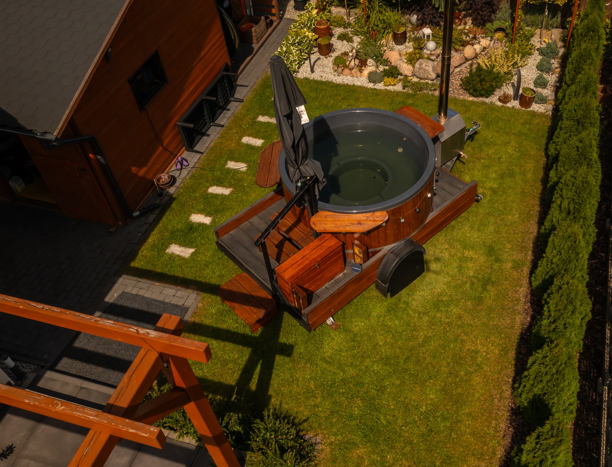 Widok z góry na ogrodową balię z wodą, z drewnianą obudową, schodkami i parasolem, umieszczoną na trawniku obok drewnianego domku i ozdobnego skalniaka.