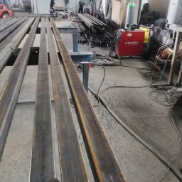 WOBISTAL WB SP. Z O.O. - Metalowe profile zamknięte, przyspawane do blach z otworami, ułożone równolegle w warsztacie, w tle spawarka Bester i pracujący ludzie.