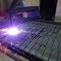 WOBISTAL WB SP. Z O.O. - Proces cięcia plazmowego arkusza metalu na stole roboczym, iskry i intensywne światło w miejscu cięcia, widoczny wzór wyciętych elementów.