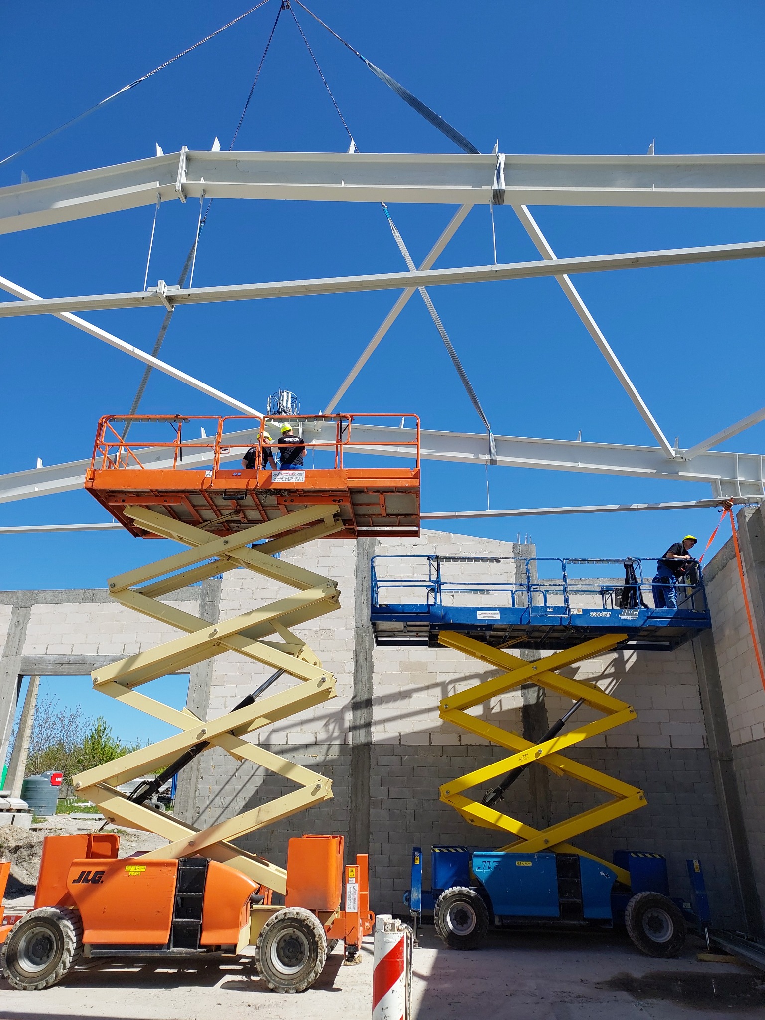 Montaż stalowej konstrukcji hali z użyciem podnośników nożycowych na tle błękitnego nieba. Dwie osoby pracują na pomarańczowym i niebieskim podnośniku.