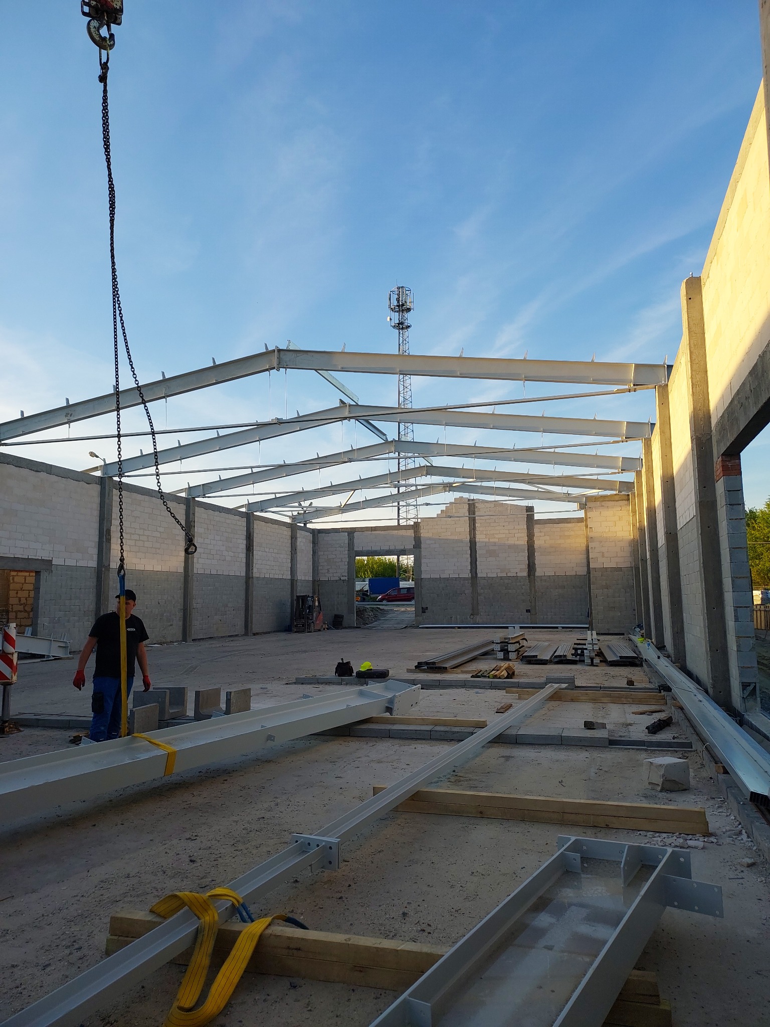 Wnętrze wznoszonej hali z bloków betonowych i stalowej konstrukcji szkieletowej, podnoszonej przez dźwig z łańcuchem. Na podłożu leżą elementy konstrukcyjne zabezpieczone żółtymi pasami.