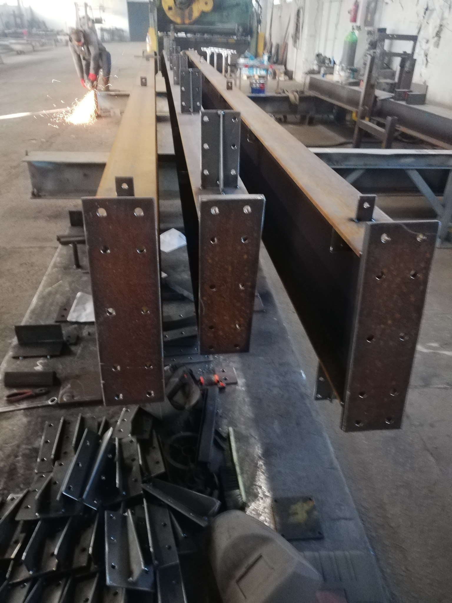 Spawanie konstrukcji stalowej hali: widok z bliska na stalowe belki z przyspawanymi płytami montażowymi; w tle spawacz w trakcie pracy z iskrami.