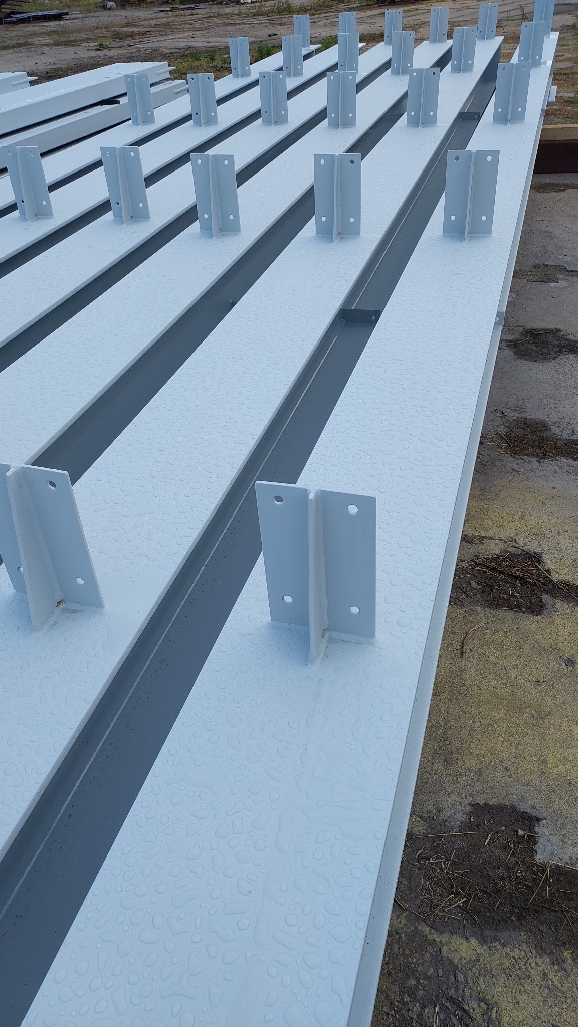 Białe, stalowe belki konstrukcyjne z przyspawanymi wspornikami, pokryte kroplami wody, przygotowane do montażu.