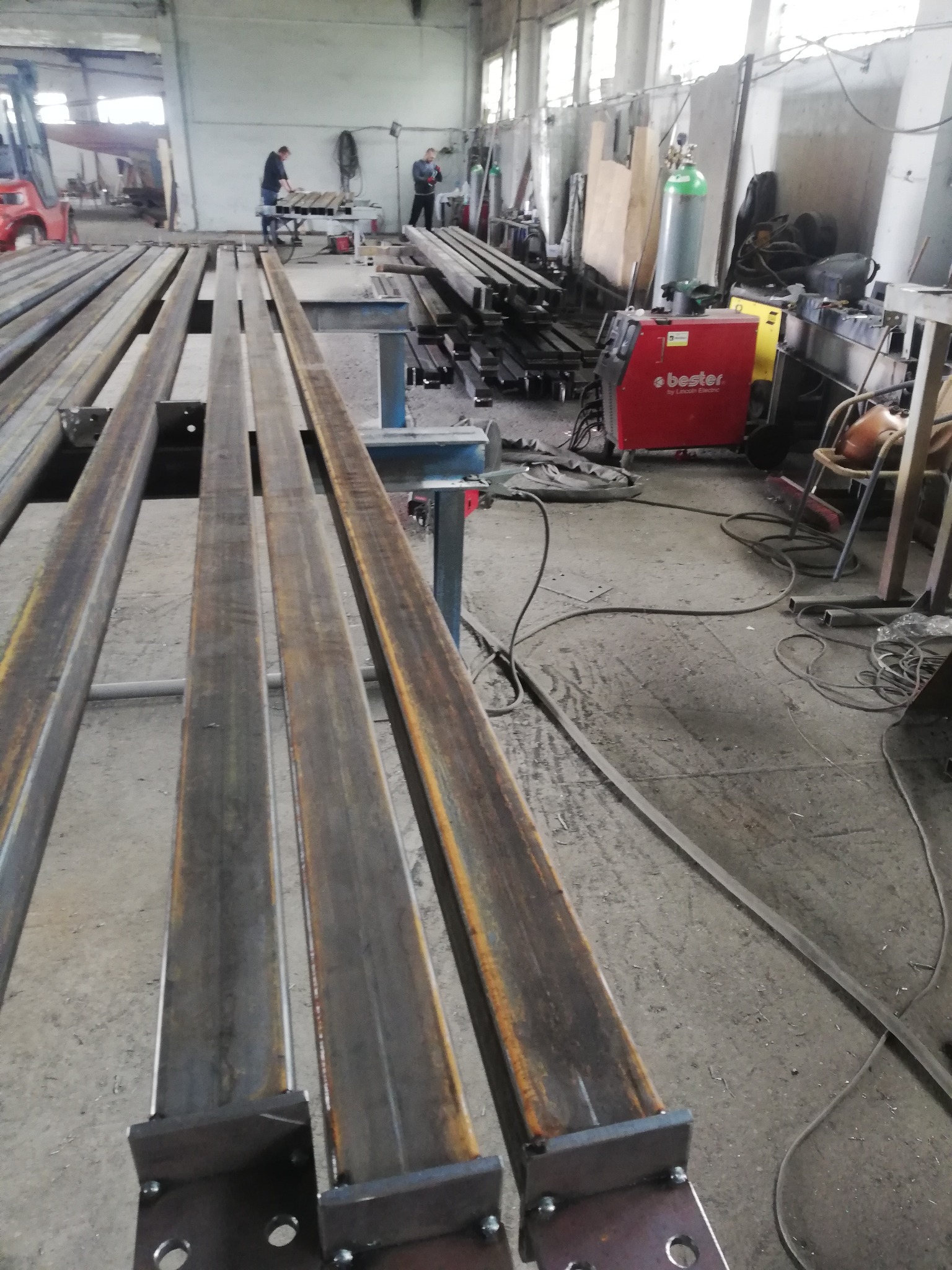 Metalowe profile zamknięte, przyspawane do blach z otworami, ułożone równolegle w warsztacie, w tle spawarka Bester i pracujący ludzie.