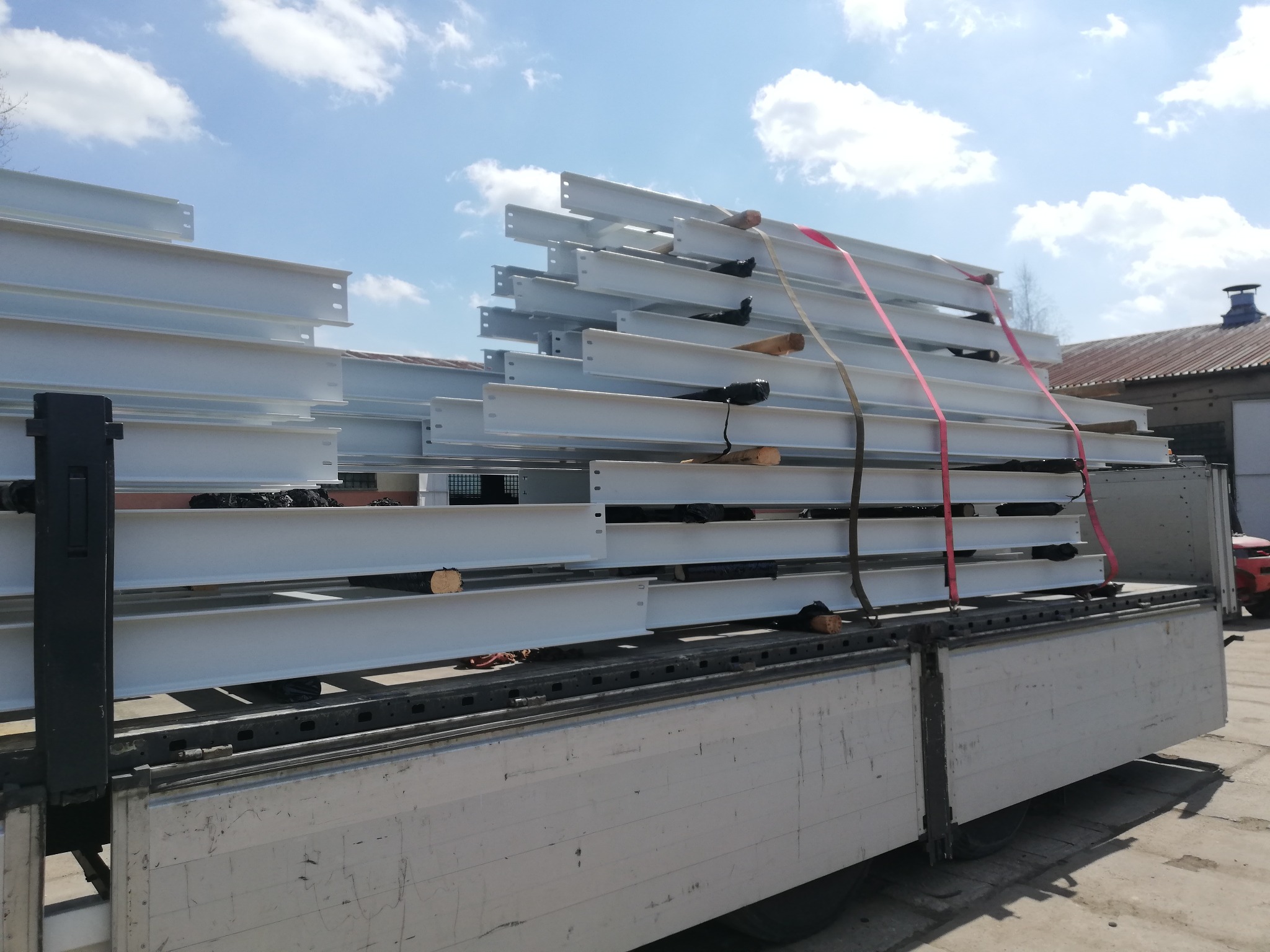 Białe, stalowe belki konstrukcyjne, zabezpieczone pasami transportowymi, ułożone na platformie ciężarówki, gotowe do przewozu na plac budowy.