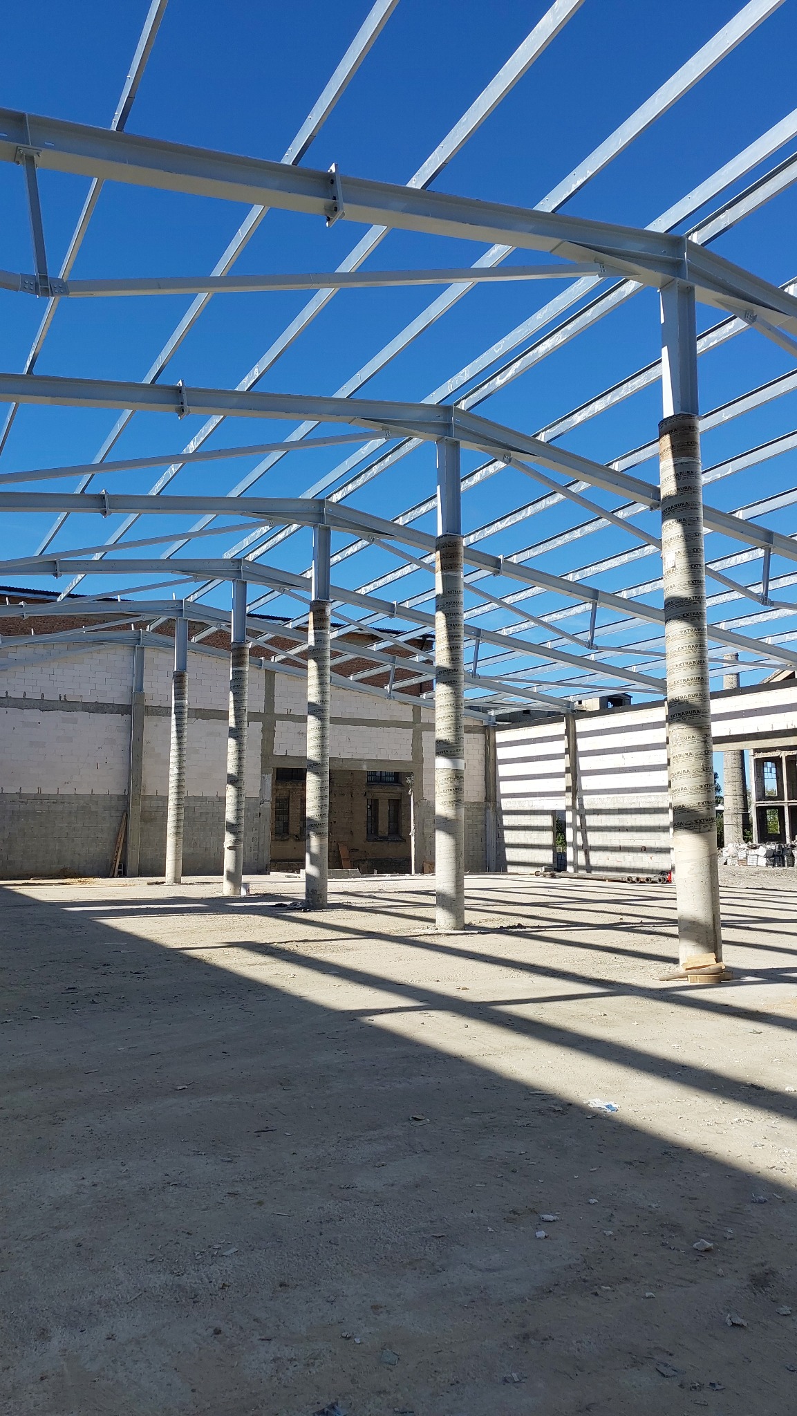 Wnętrze hali w trakcie budowy, widoczne stalowe elementy konstrukcyjne i betonowe słupy podtrzymujące, oświetlone jasnym światłem słonecznym tworzącym wyraźne cienie na betonowej podłodze.