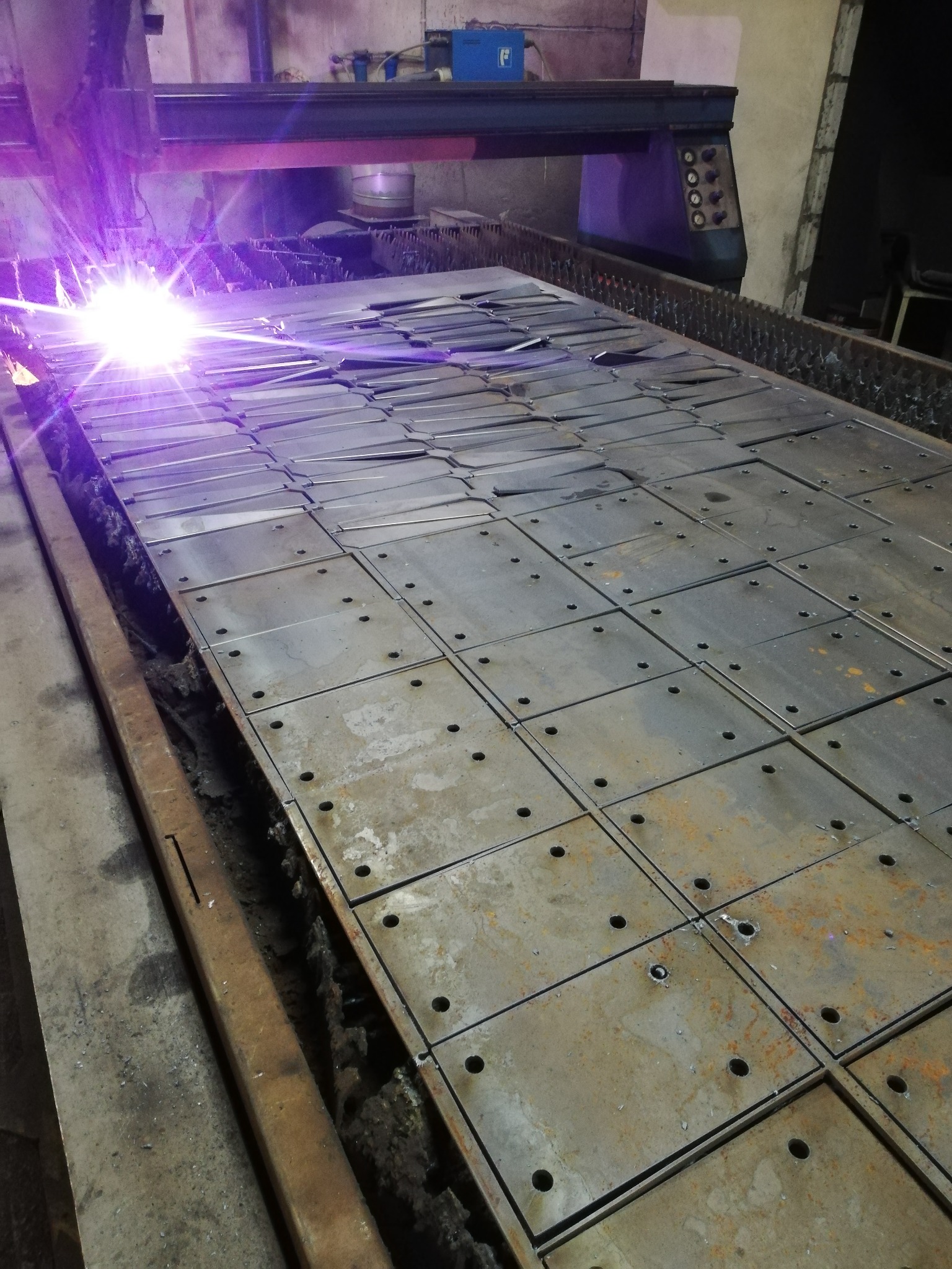 Automatyczna wycinarka plazmowa tnie arkusz blachy na kwadratowe elementy z otworami, widoczne iskry i ciemne otoczenie hali produkcyjnej.