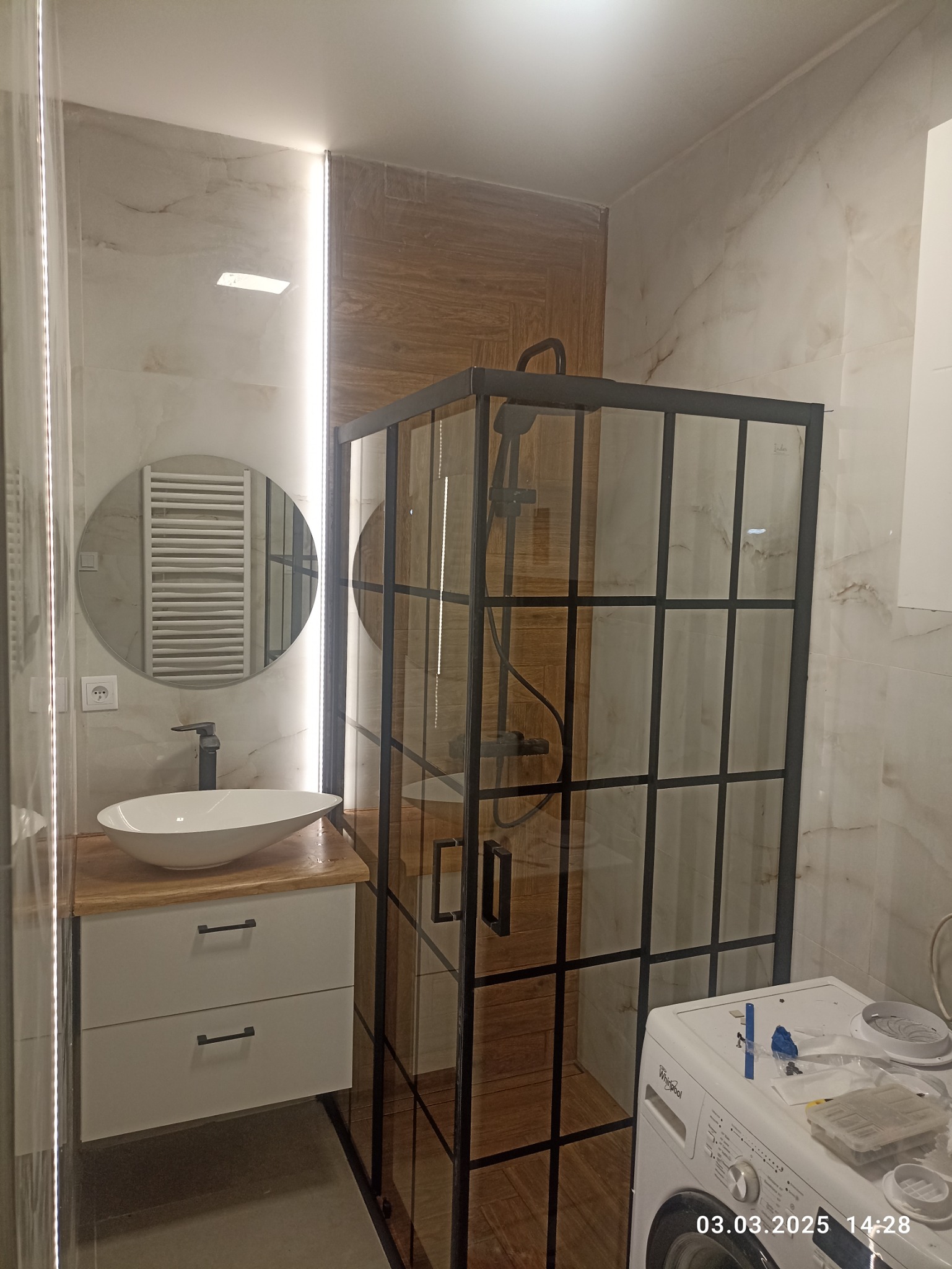 Nowoczesna łazienka z kabiną prysznicową typu loft, umywalką nablatową na drewnianym blacie i okrągłym lustrem z podświetleniem LED. Marmurowe płytki na ścianach.