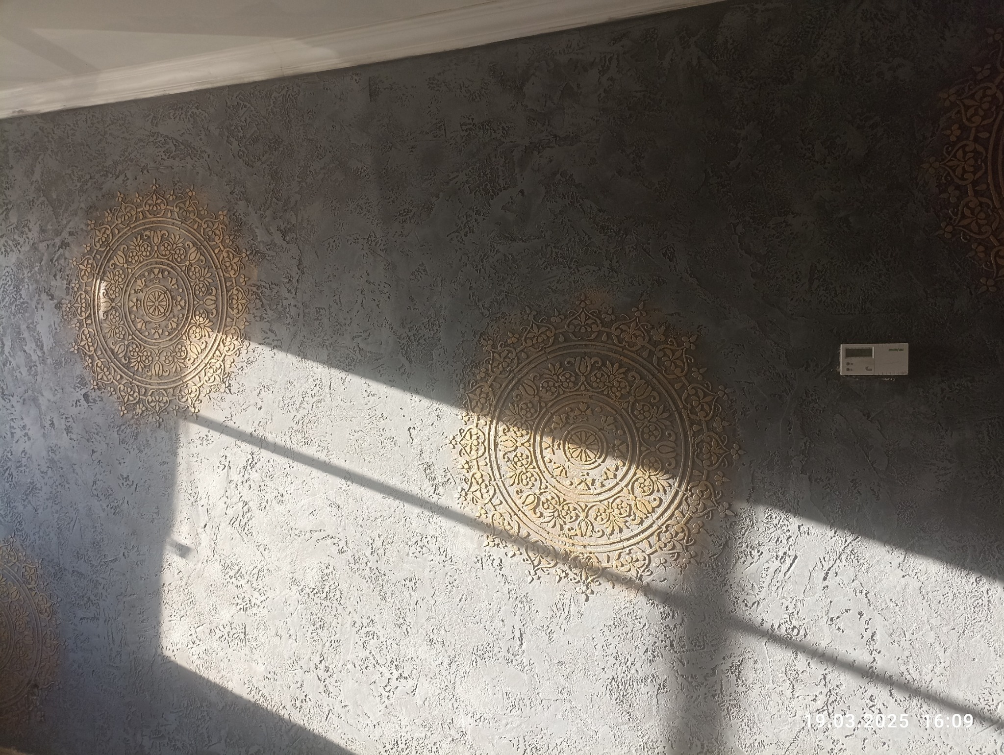 Dekoracyjna ściana z efektem stiuku w odcieniach szarości i złota, zdobiona ornamentami w stylu mandali. Wykończenie wnętrz w Pile.