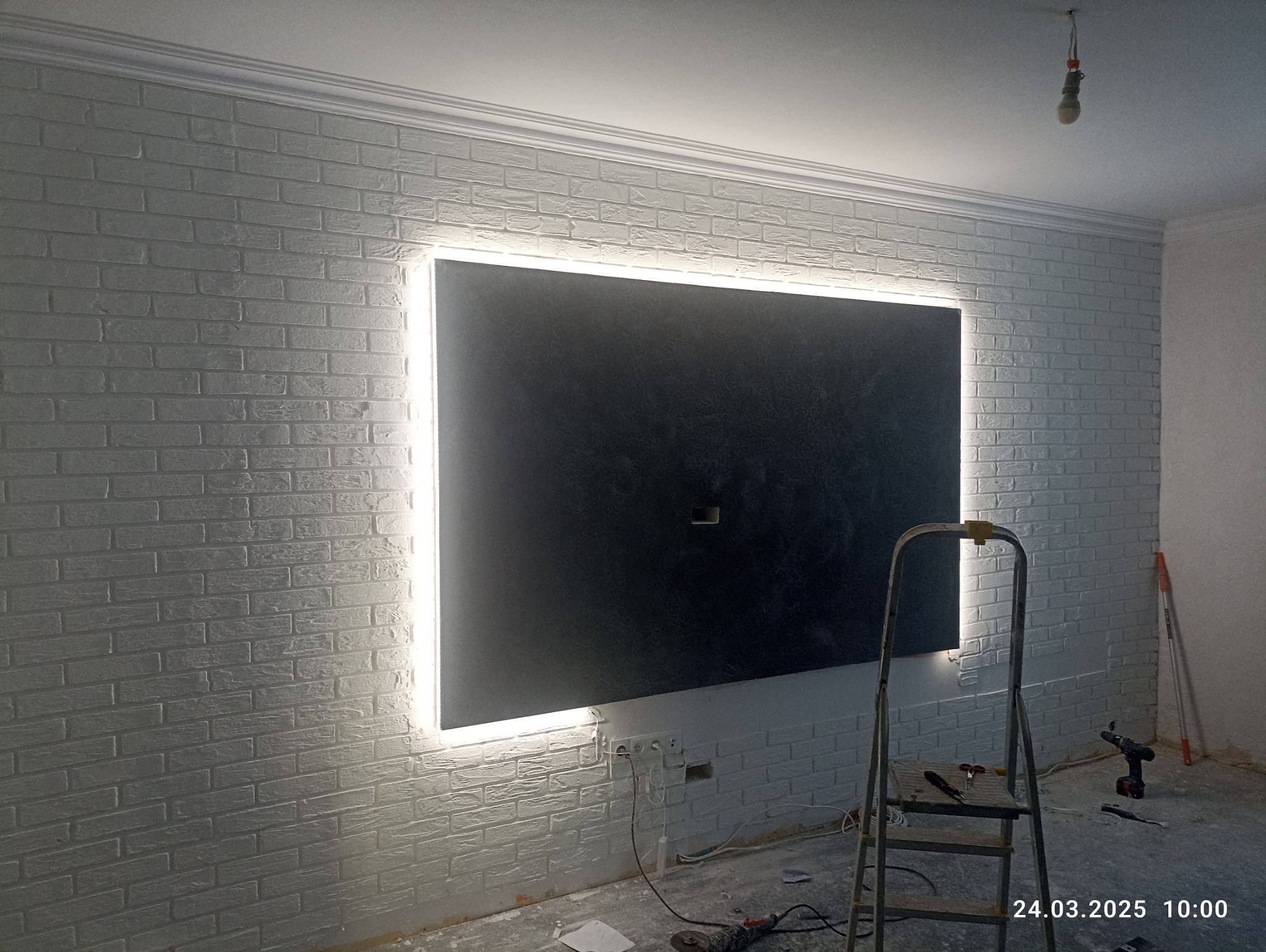 Ściana z białej cegły z podświetlonym panelem dekoracyjnym. Widoczna drabina i narzędzia, sugerujące prace wykończeniowe w trakcie realizacji.