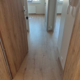 Sebastian Gajewski - Widok na wnętrze mieszkania z perspektywy otwartych drzwi wejściowych, ukazujący panele podłogowe w odcieniach drewna, białe ściany oraz fragment maty podłogowej przy progu.