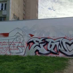 Sebastian Gajewski - Biała ściana z graffiti przedstawiającym częściowo zamalowany szkic postaci z pędzlem oraz kolorowy napis w stylu graffiti, z tekstem po polsku napisanym czerwoną farbą, widok z poziomu trawnika.