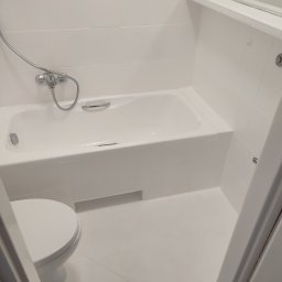 Sebastian Gajewski - Całkowicie biała łazienka z wanną, toaletą i białymi kafelkami, świeżo po remoncie, widoczne ślady farby na podłodze.