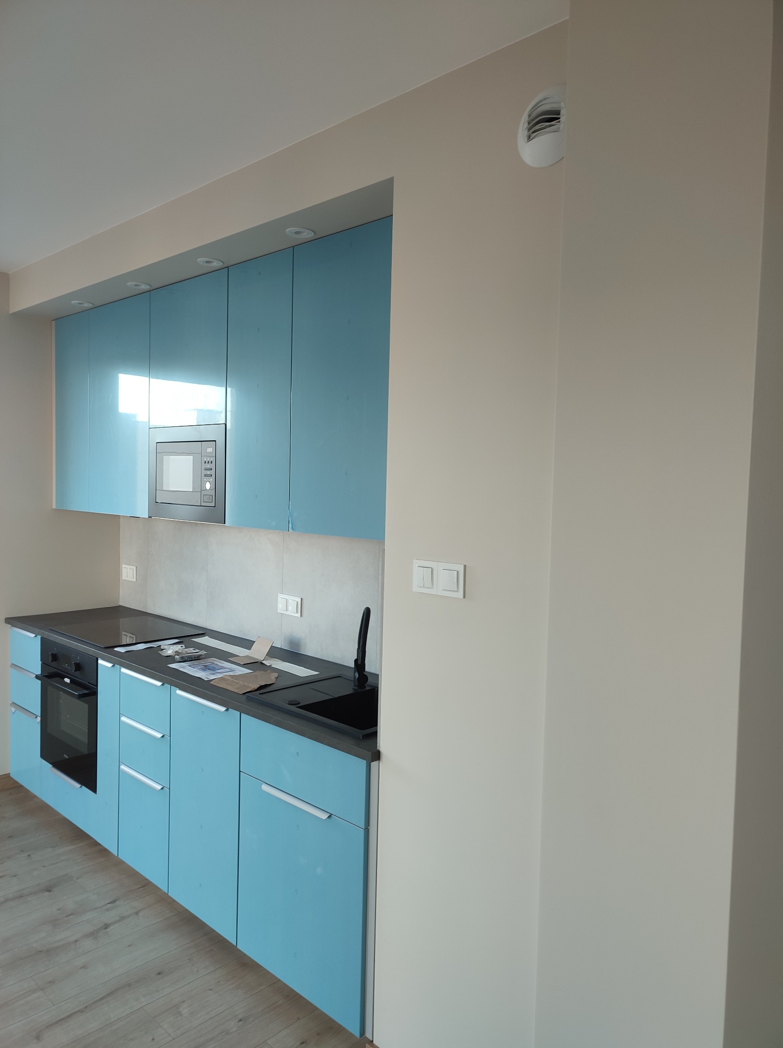 Niebieskie, lakierowane szafki kuchenne z szarym blatem i sprzętem AGD, widoczne detale wykończenia ścian i podłogi.