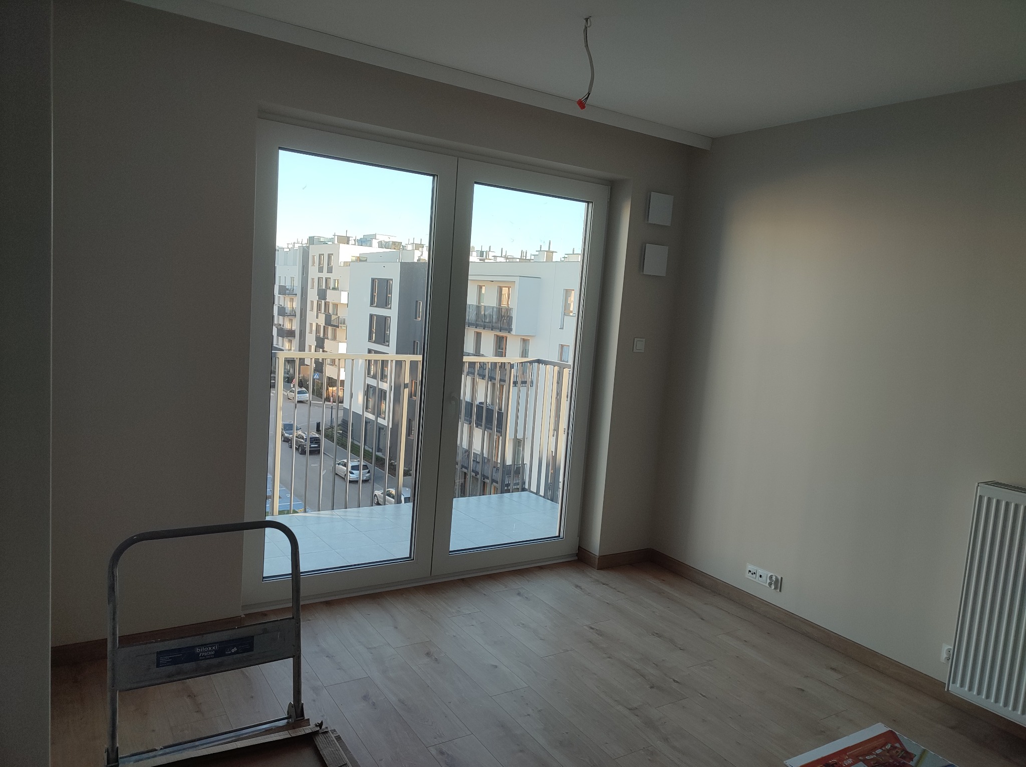 Pomalowane na jasny beżowo-szary kolor ściany w pustym pokoju z drewnianą podłogą, widok na osiedle przez dwuskrzydłowe drzwi balkonowe, drabina malarska w rogu.