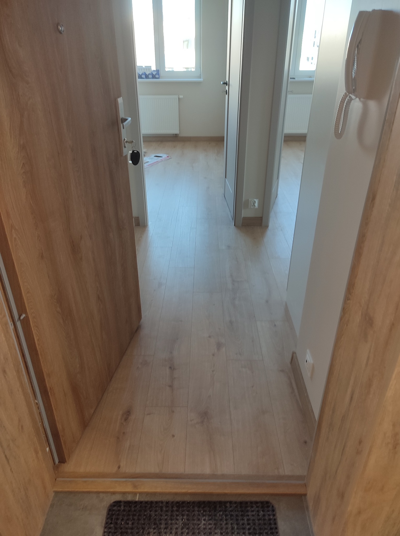 Widok na wnętrze mieszkania z perspektywy otwartych drzwi wejściowych, ukazujący panele podłogowe w odcieniach drewna, białe ściany oraz fragment maty podłogowej przy progu.