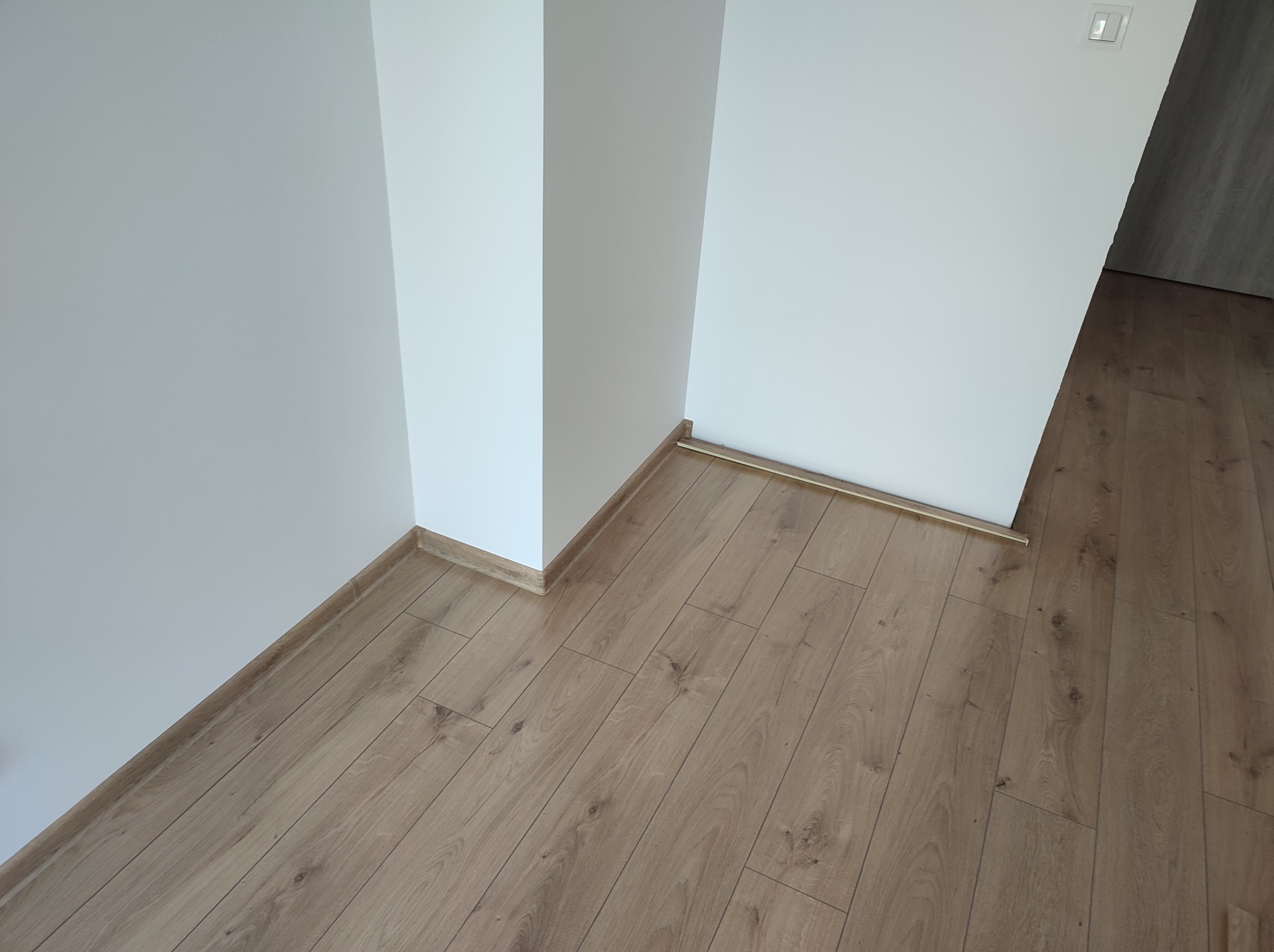 Jasne panele podłogowe ułożone ukośnie w rogu pomieszczenia z białymi ścianami i listwami przypodłogowymi w kolorze podłogi, widoczny włącznik światła.