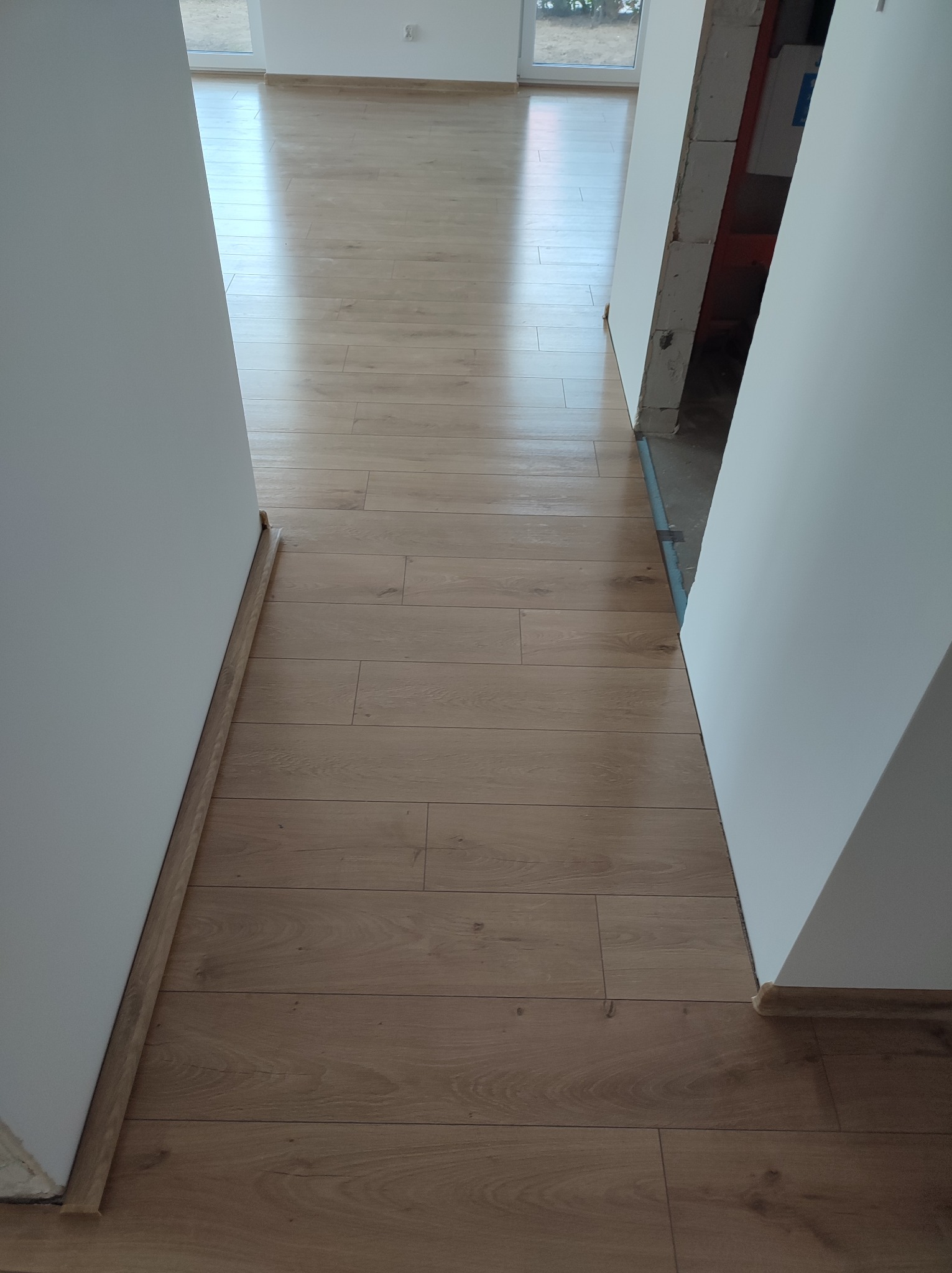Ułożona, jasna podłoga laminowana w korytarzu, widoczne białe ściany i fragment surowego otworu drzwiowego z widocznym betonem i instalacjami.