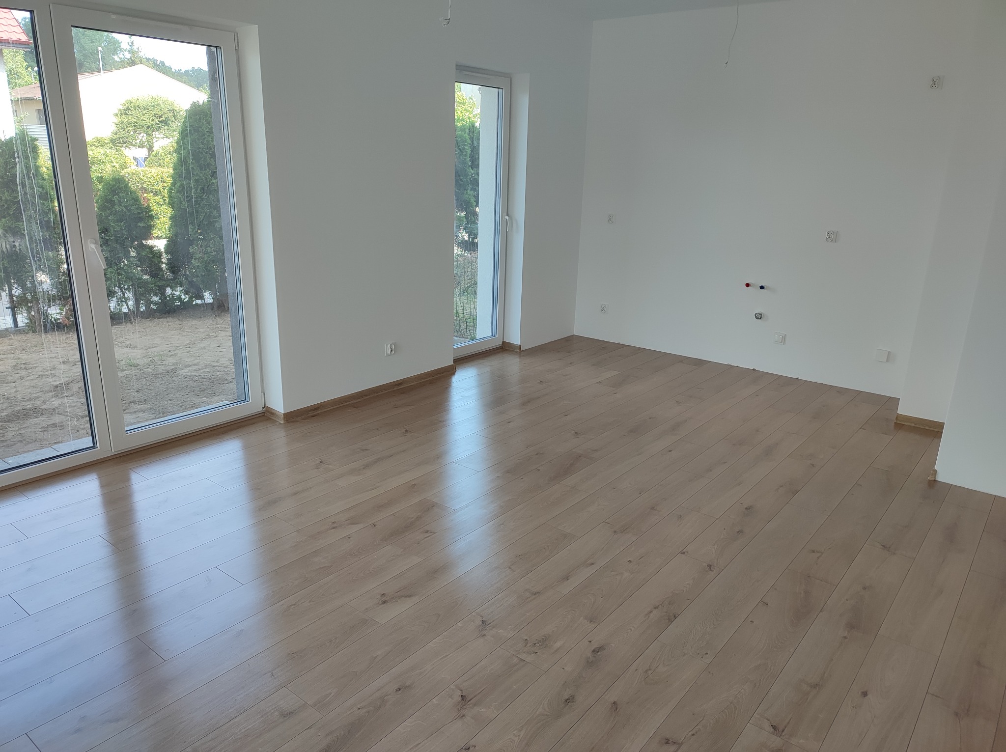 Jasne panele podłogowe w pustym, świeżo odnowionym pokoju z białymi ścianami i dwoma oknami wychodzącymi na zieleń.